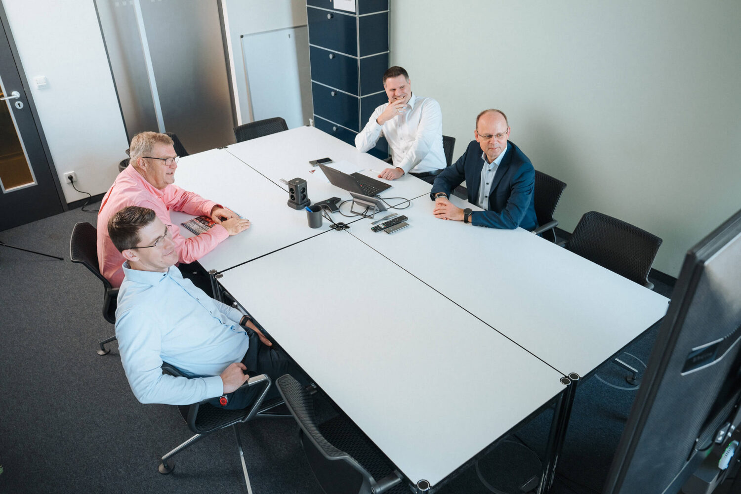 Vier Personen sitzen an einem weißen Konferenztisch in einem Konferenzraum und blicken auf einen Bildschirm