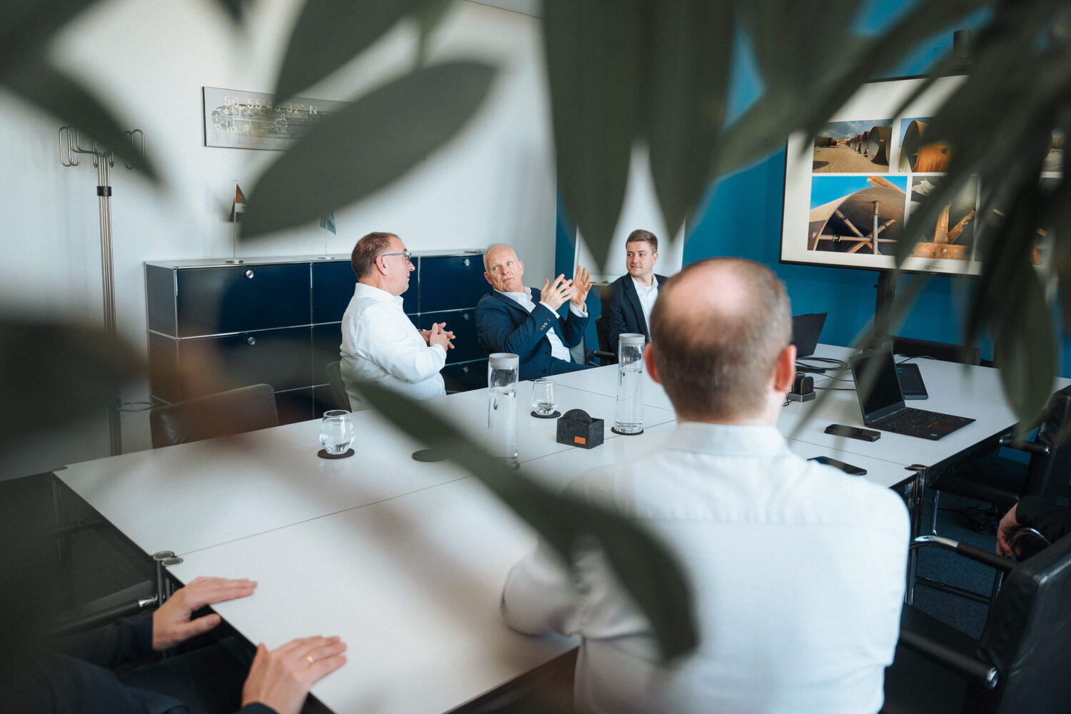 Mehrere Mitarbeiter sitzen an einem Konferenztisch in einem Büro, im Vordergrund sind Blätter unscharf zu sehen